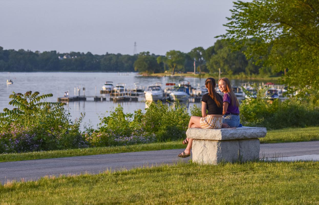 Friends catching up on a bench at Perrysburg Riverside Park with water and docked boats in the distance. 