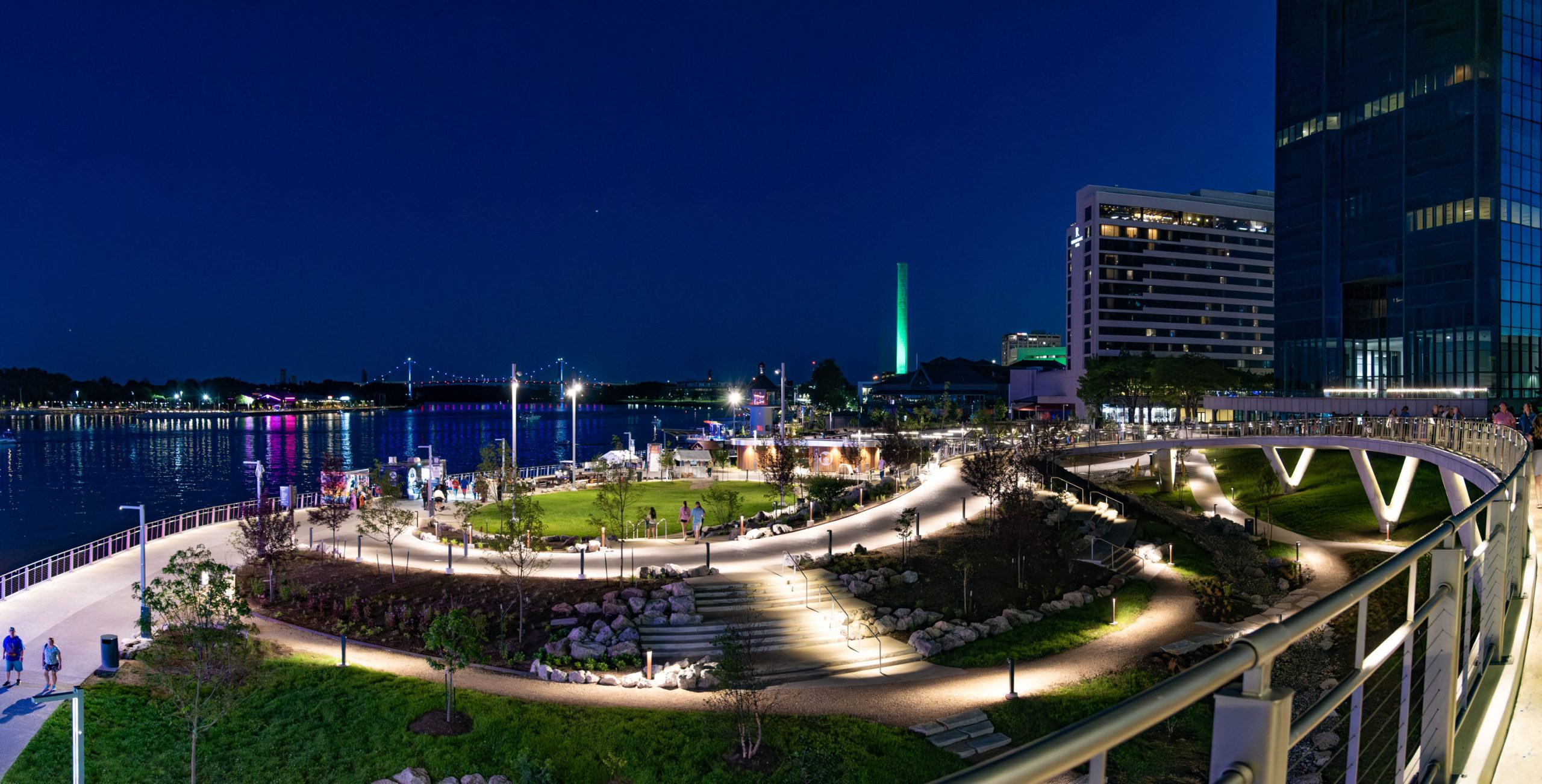 Glass City Riverwalk, Toledo Ohio. Night scene from MLK Bridge to Basin event lawn during event. People walk down bridge ramp toward concentric oval shaped lawn with elevation change, stairs, glowing lights illuminate pathways and uplighting illuminates bridge and plant material. In the background, the bridge and river glow with colorful lights and reflections.